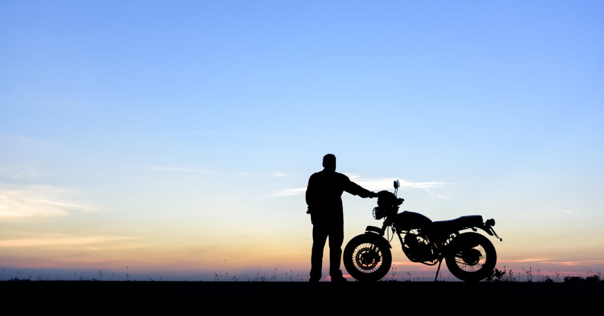 A person stands in silhouette beside a motorcycle against an orange sunset sky. The person gazes into the distance.