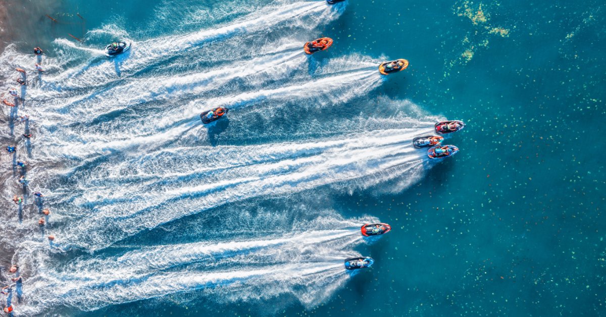 An aerial view of multiple personal watercraft racing across blue water with curved wakes trailing behind.