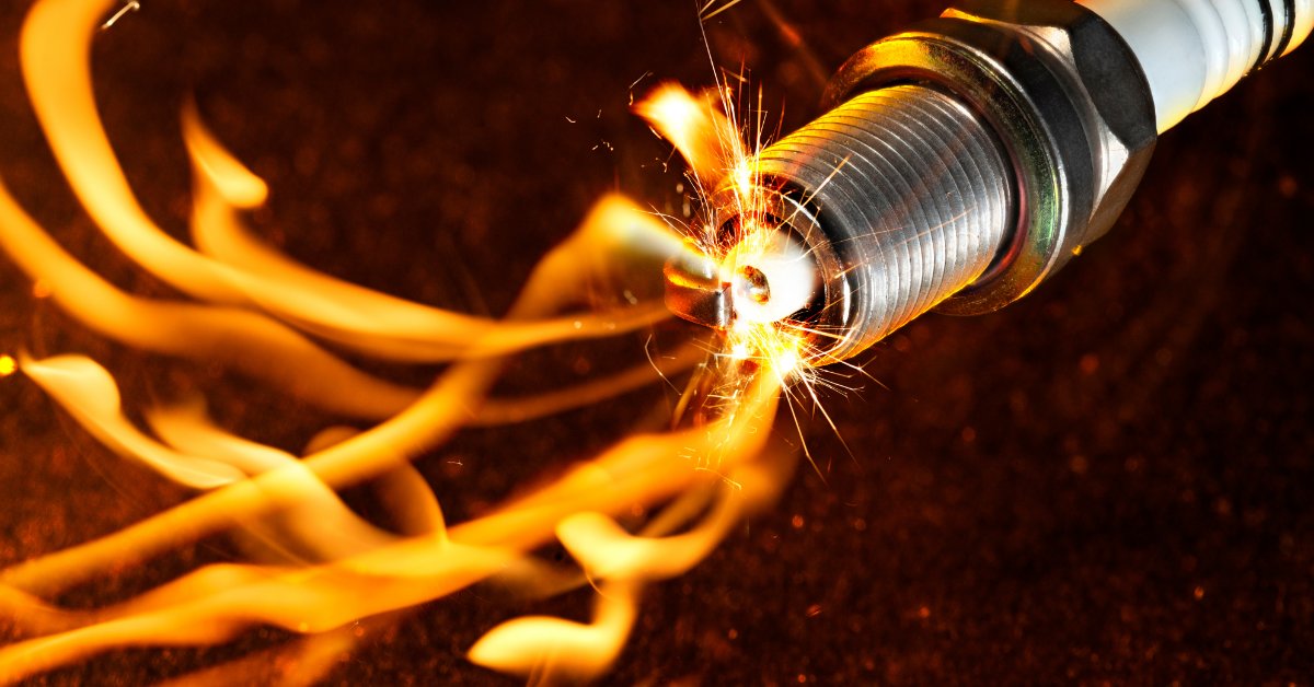 A metal spark plug with a white ceramic insulator producing bright orange sparks amid flames against a dark background.