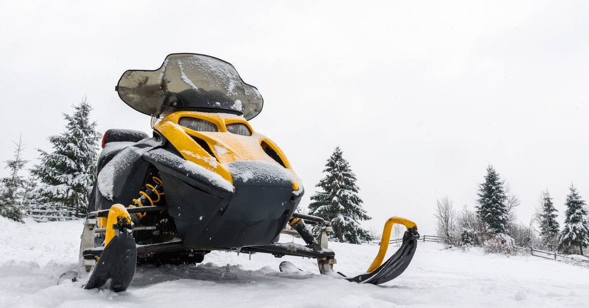 A front view of a yellow snowmobile parked on snowy terrain around several pine trees in a fenced area.