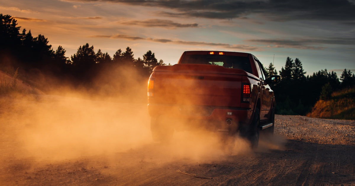 A pickup truck driving on a country road, with dust trailing behind it as it heads toward a forest landscape.