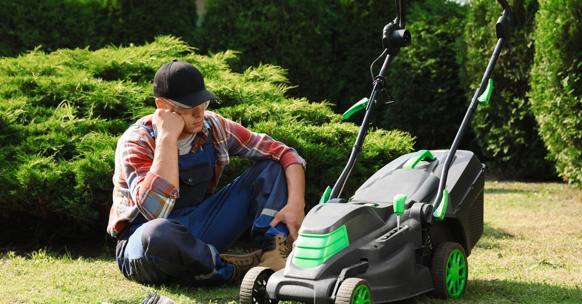 A young man sits next to his lawn mower in a yard, looking defeated. Beside him lie a screwdriver and a brush.