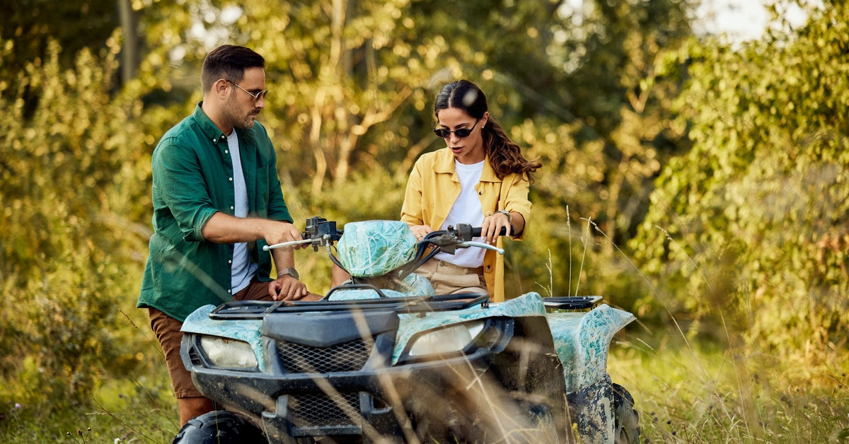 A man and woman inspect their quad bike while exploring a forested area. They grip the handles as they examine it.