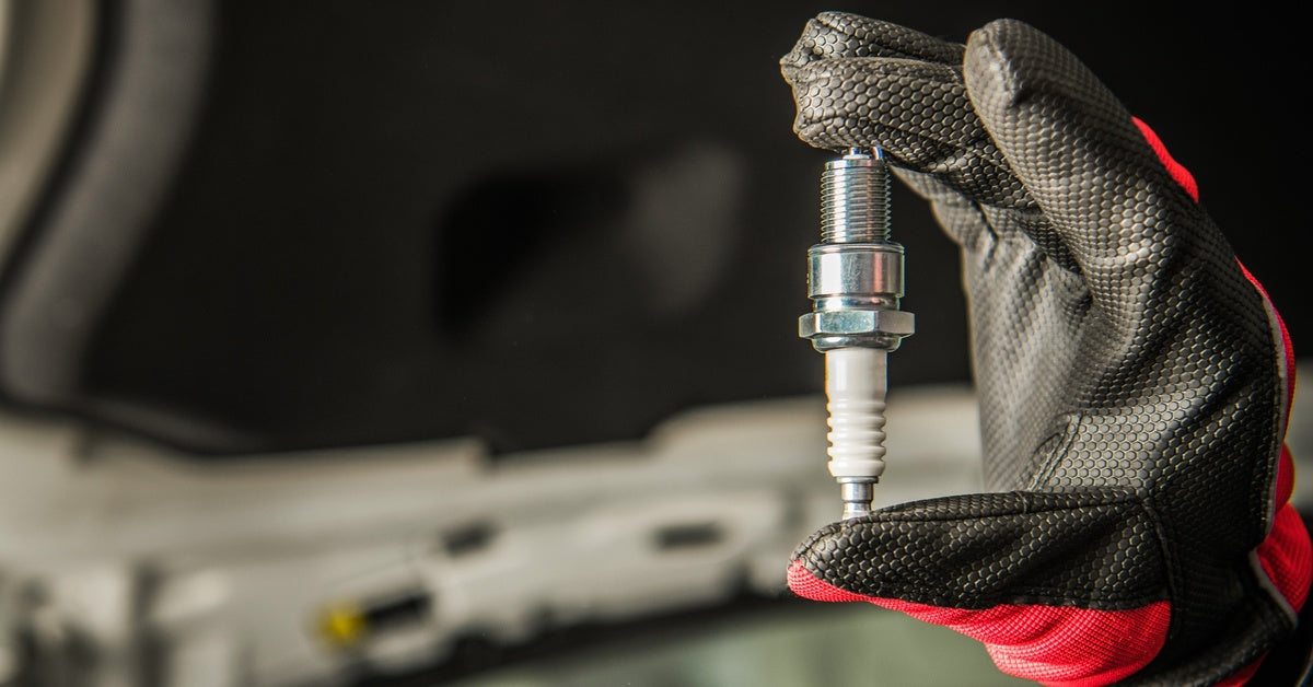 A hand wearing a black and red leather glove holds a clean spark plug up against a blurry background.