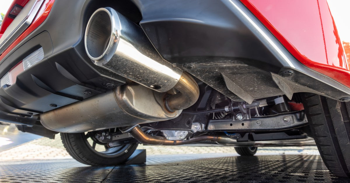 Low-angle view of a red car's underside, showing a shiny chrome exhaust pipe, muffler, and dark underbody components.