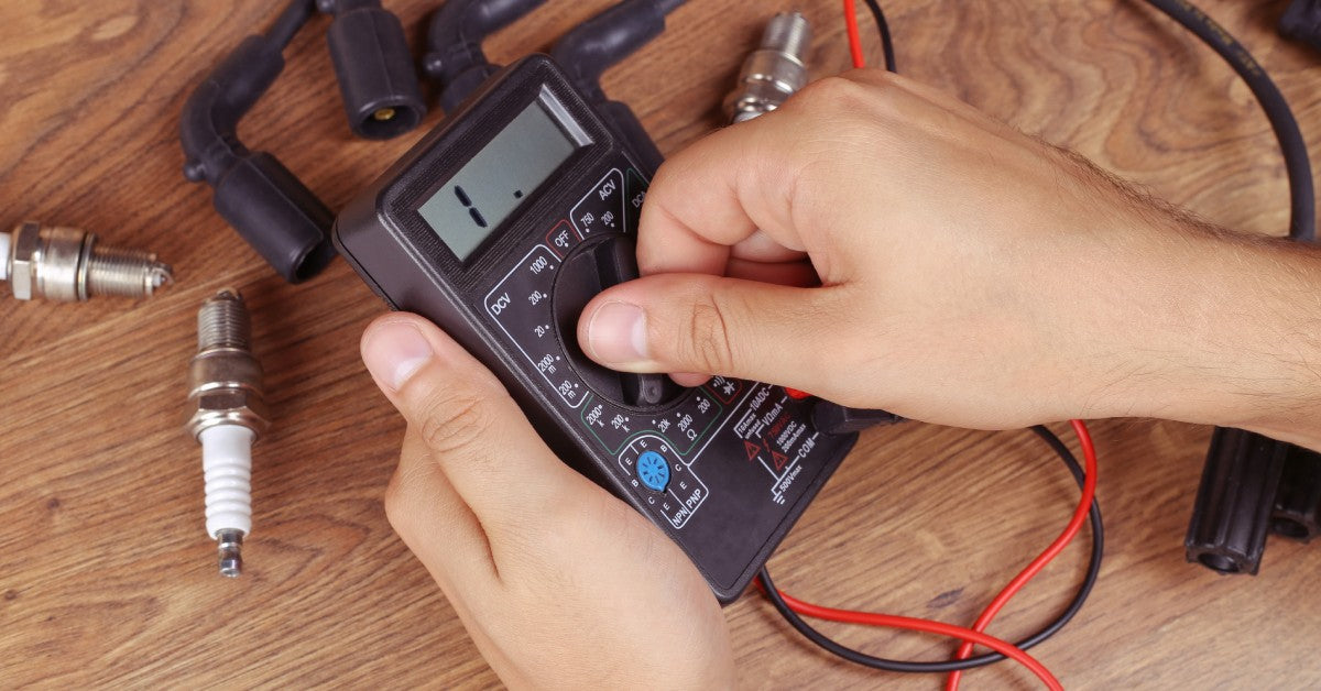 A person holds an electrical test kit, with two spark plugs and other cables arranged on a wooden surface.