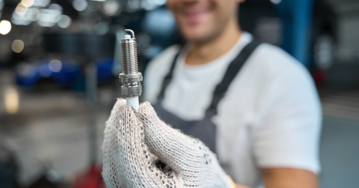 A gloved hand holds a spark plug, set against the blurred background of a smiling man in a garage-like setting.