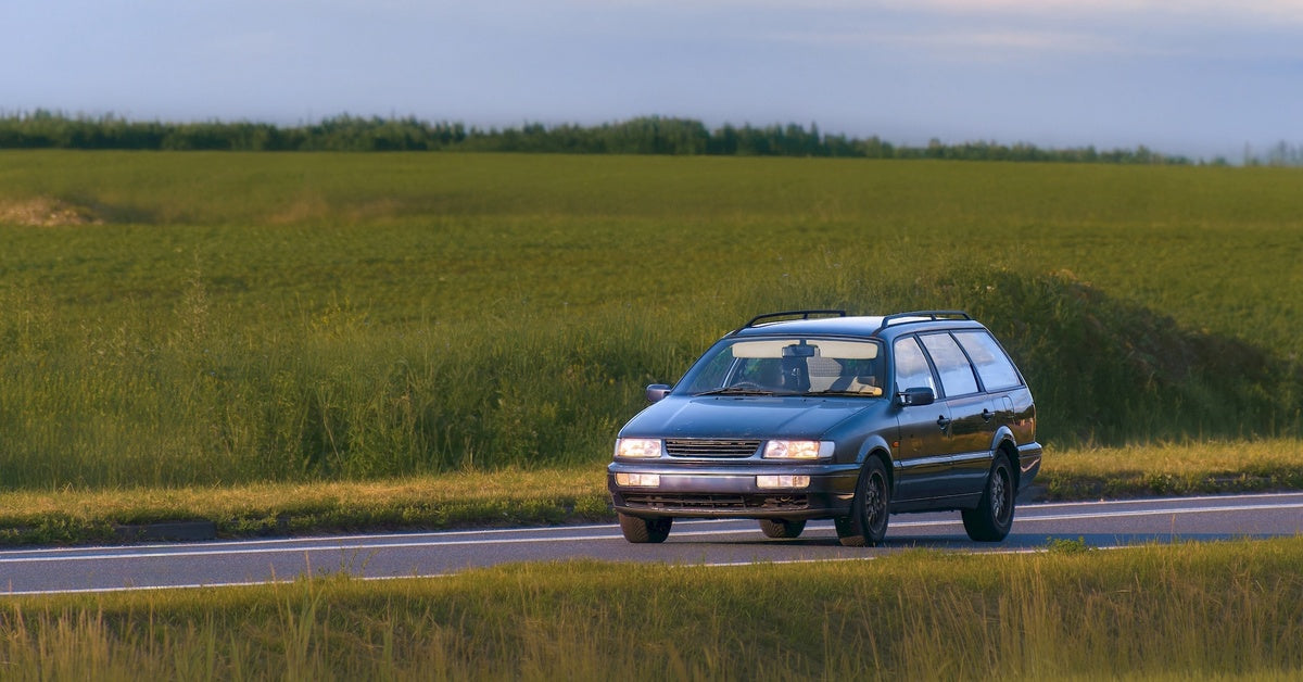 A dark blue, couple-style car drives along an open road between fields of green grass under a sunny sky.