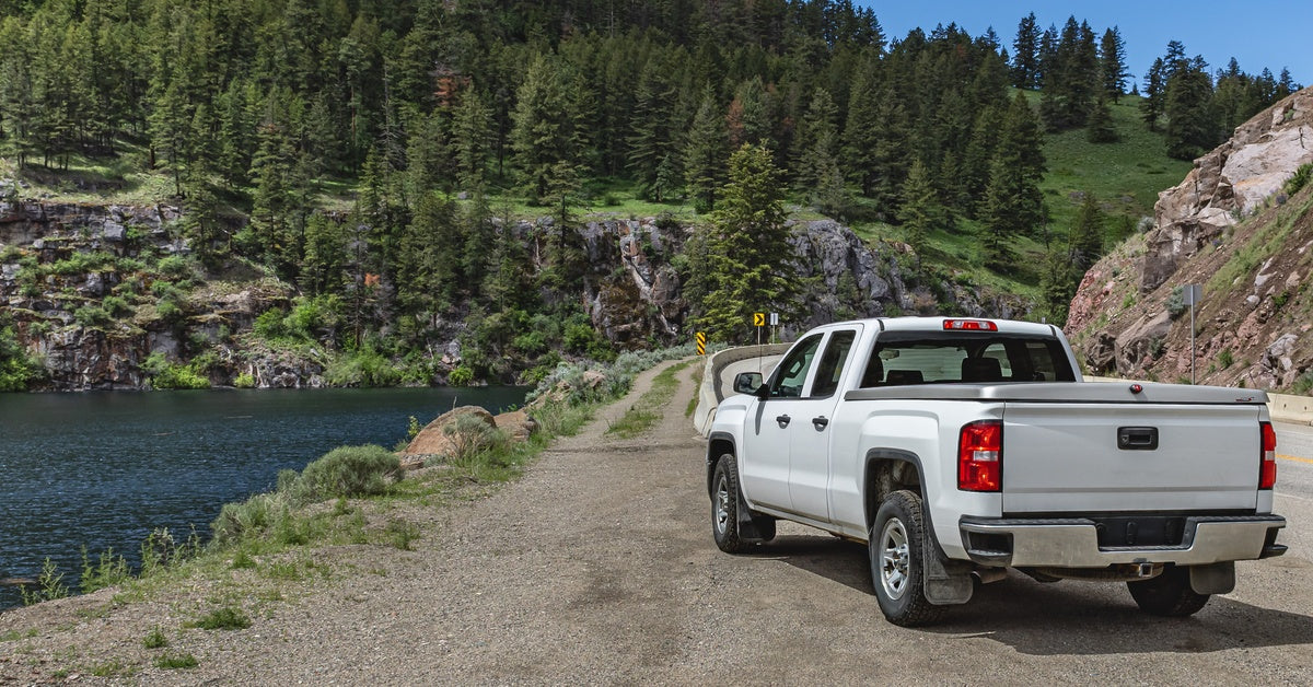 White pickup truck parked beside a mountain lake on a dirt road, surrounded by pine trees and rocky cliffs.