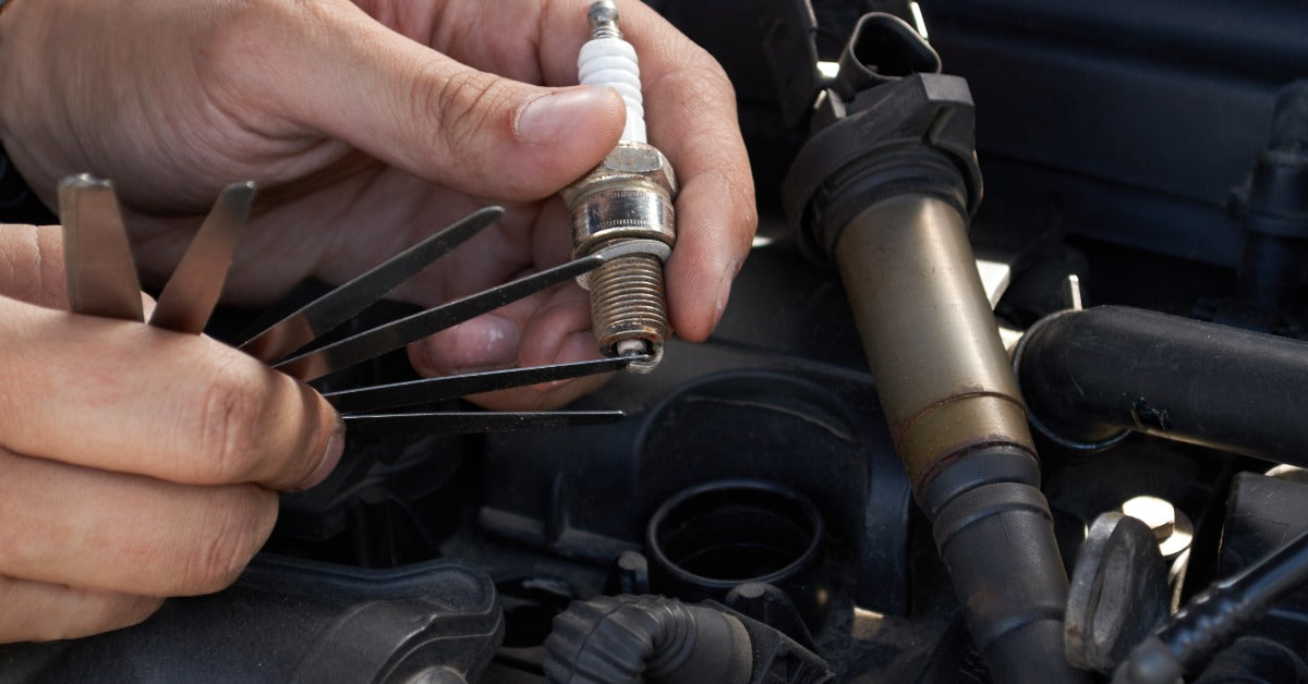 A close-up on a pair of hands using a feeler gauge to measure spark plug gap above an open car engine bay.