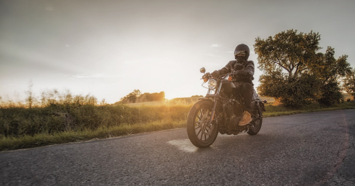 Motorcyclist riding a cruiser on an open road during golden hour sunset, with trees and countryside landscape visible.