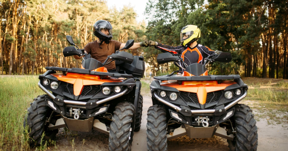 Two riders with helmets seated in parked ATVs on a muddy forest trail exchange a fist bump between the vehicles.