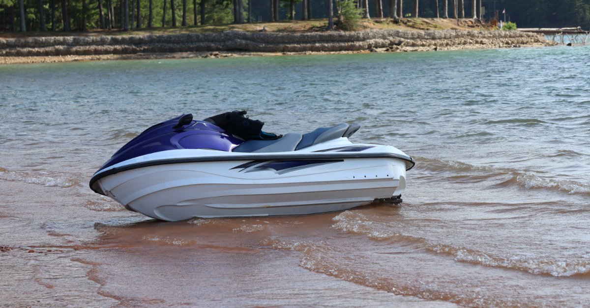 A blue and white jet ski is parked beside a sandy shore with calm water and a shoreline in the background.