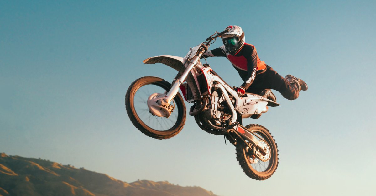 A motocross rider on a white dirt bike performs a jump against a clear sky with the front wheel raised.