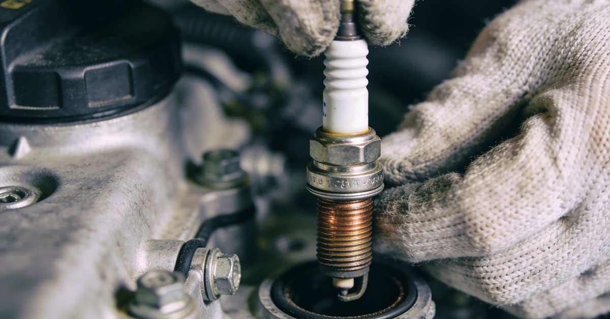 A close-up of a person removing a spark plug with dirty threads from inside an open car engine bay with gloves.