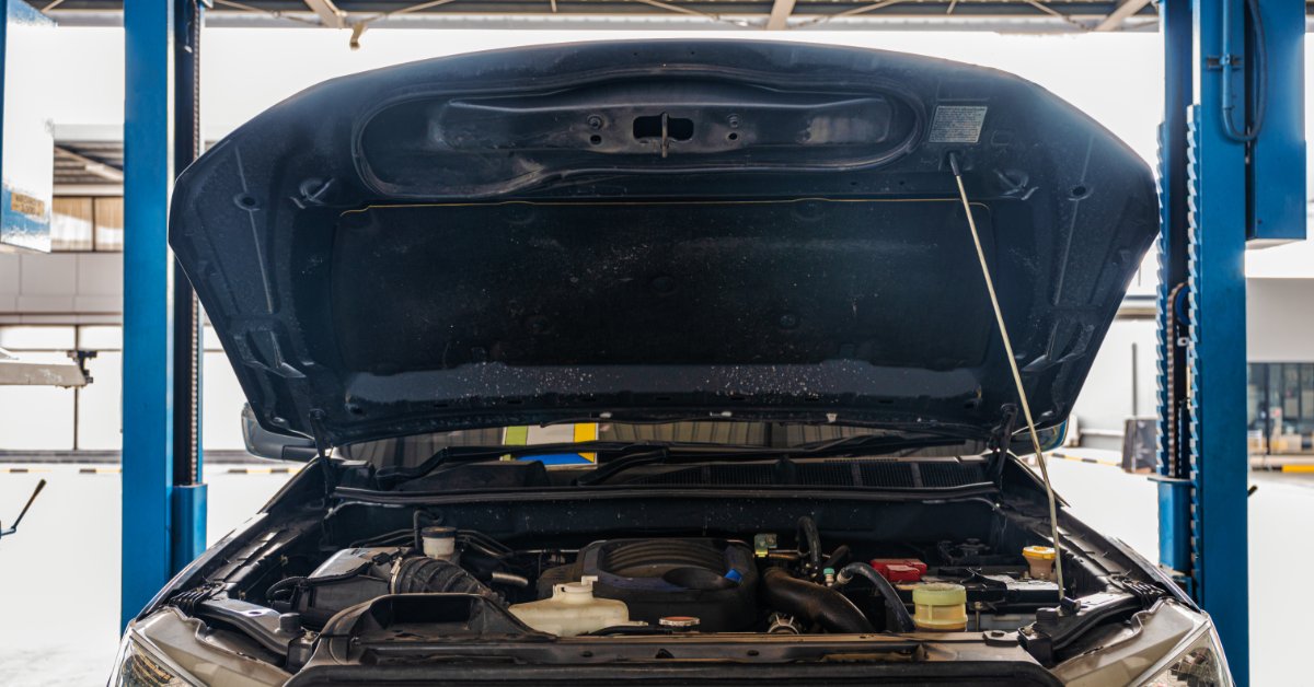 A pickup truck with its hood open inside a repair garage, showing the engine bay between two blue lift posts.