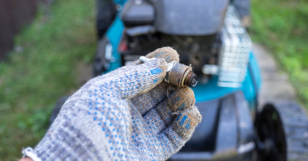 A close-up of a person with gloves holding a spark plug in front of a blue lawn mower on a grassy pathway.