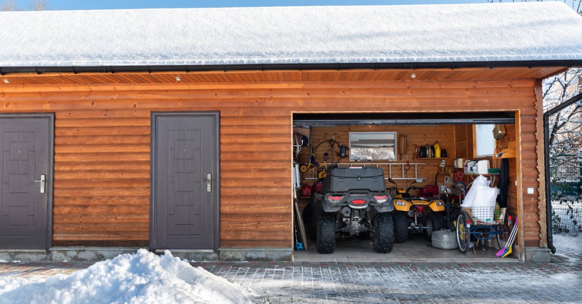 A wooden garage with snow on the roof has its doors open, showing parked ATVs, tools, and storage items.