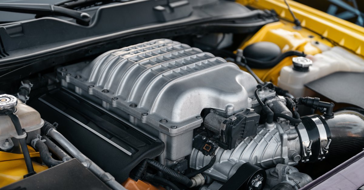 A close-up of a shiny car engine with a large metal supercharger, belts, hoses, and yellow body panels visible.