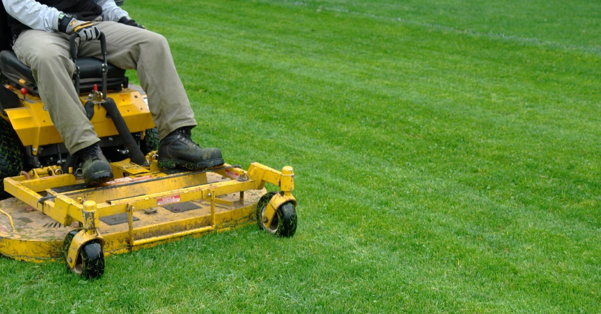 A close-up of a person riding a yellow lawn mower on trimmed grass, with an open lawn space to the right.
