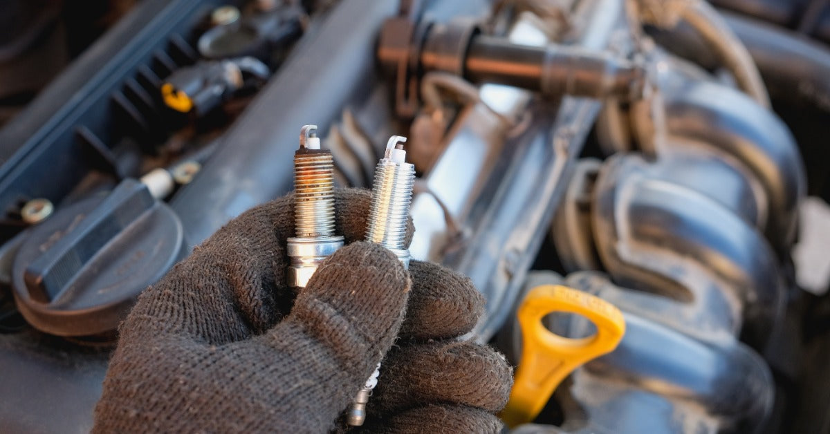 A close-up of a person holding two spark plugs, one worn and one clean, above an open car engine compartment.