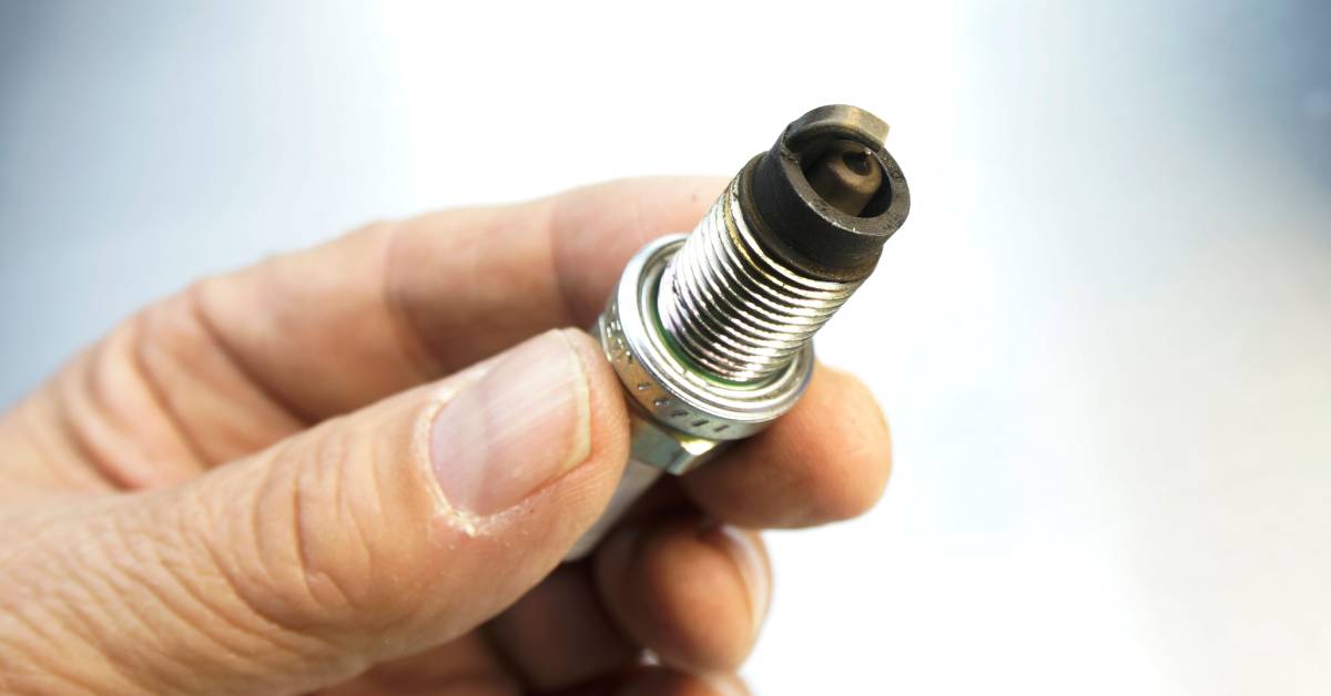 A man's hands holding a fouled automotive spark plug. The insulator of the spark plug is black and burnt.