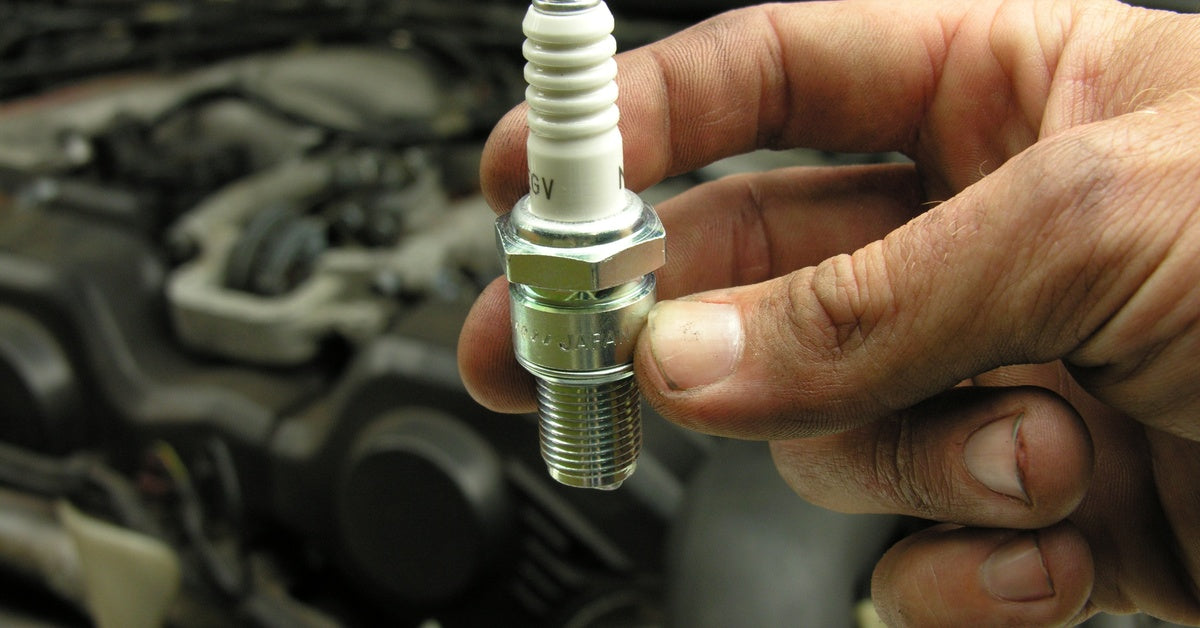 A close-up of a person holding a brand-new spark plug, with the part number and stock numbers visible.
