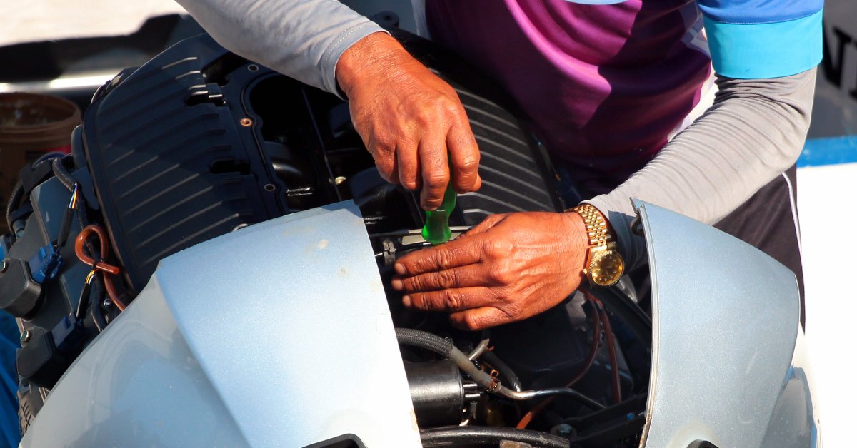 A man wearing a gold watch and a cap uses a green screwdriver to remove parts from the top of a speedboat.