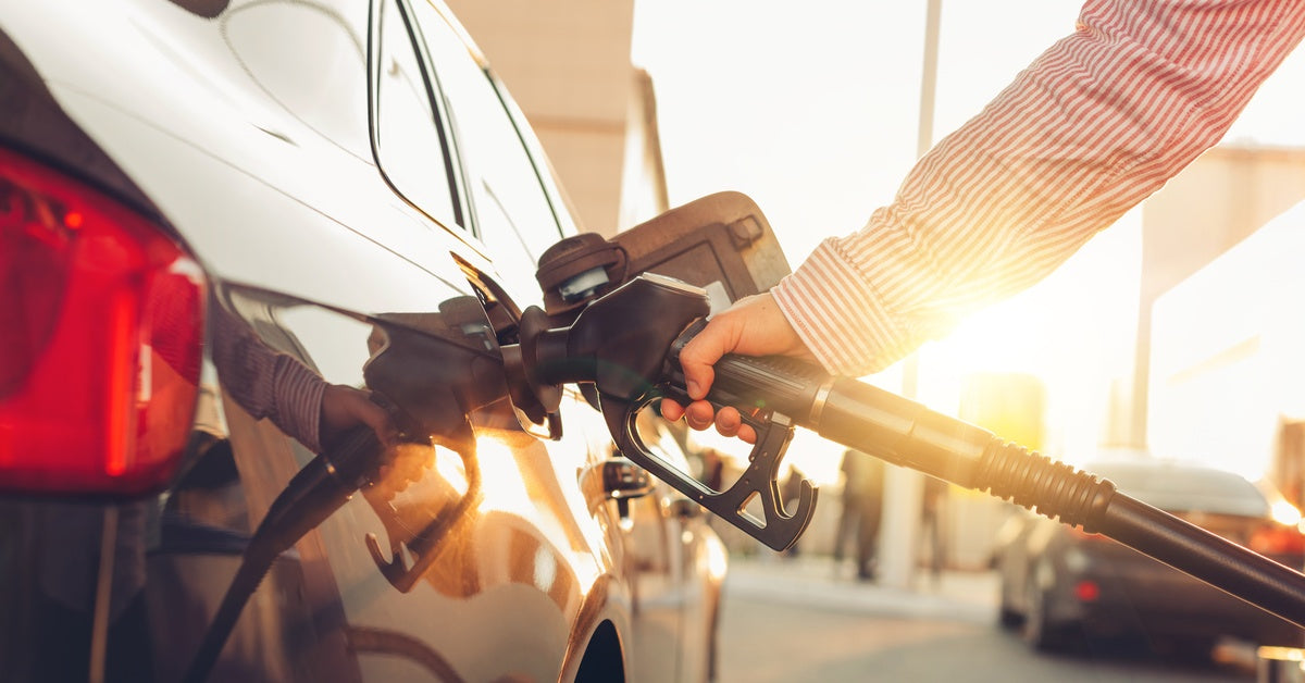 A person in a red striped long-sleeved shirt holds a fuel nozzle to refuel their black car at a gas station.