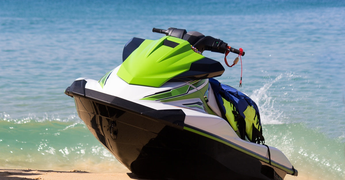 A green-and-white jet ski is parked on a sandy beach, with waves crashing against the shoreline behind it.