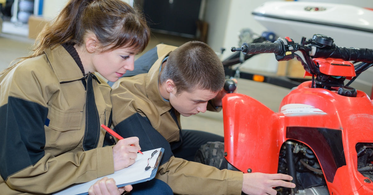 Two mechanics inspect an ATV in a garage, examining the vehicle's parts while taking notes on a clipboard.
