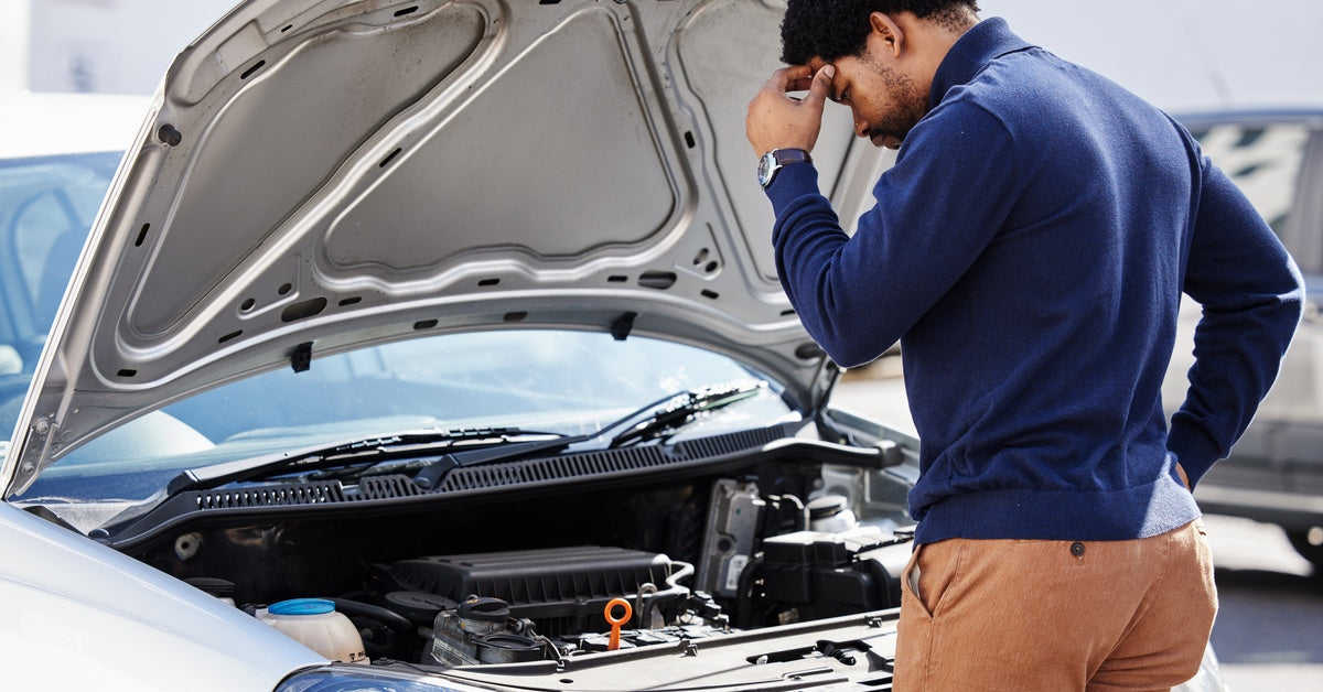 A Black man in a parking lot looks at his car engine in confusion, trying to determine what the problem is.