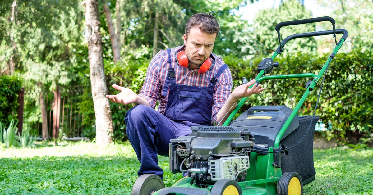 A man wearing denim overalls and noise-protecting gear looks at a green lawn mower with a frustrated expression