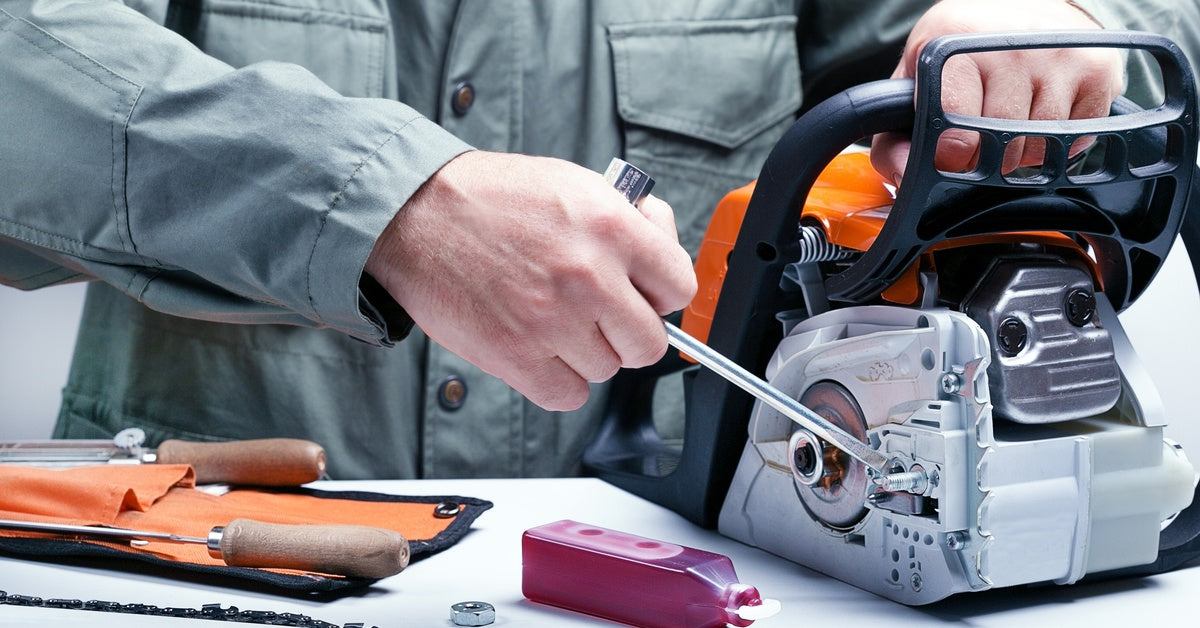 A person uses a tool to adjust a disassembled chainsaw at a workspace. There are other materials on the table.