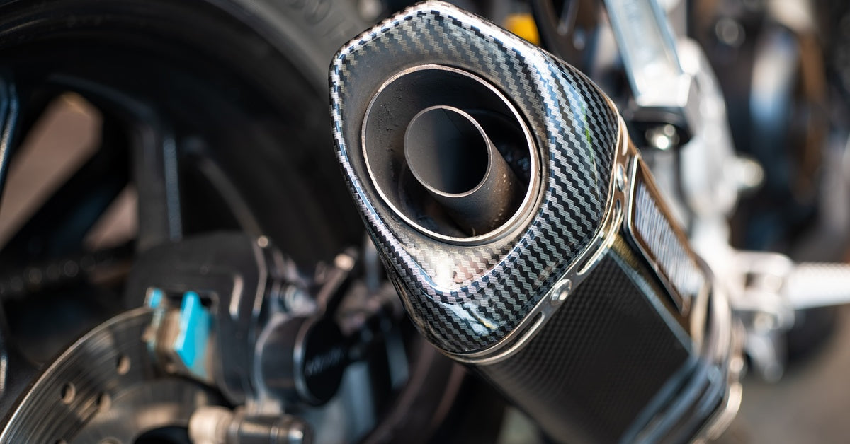 Close-up of a motorcycle exhaust with a carbon fiber pattern, silver rim, and blurred rear wheel and brake disc in view.