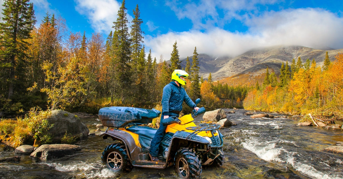 A person wearing a blue sports suit and a yellow safety helmet stands on an ATV in a small river near the mountains.