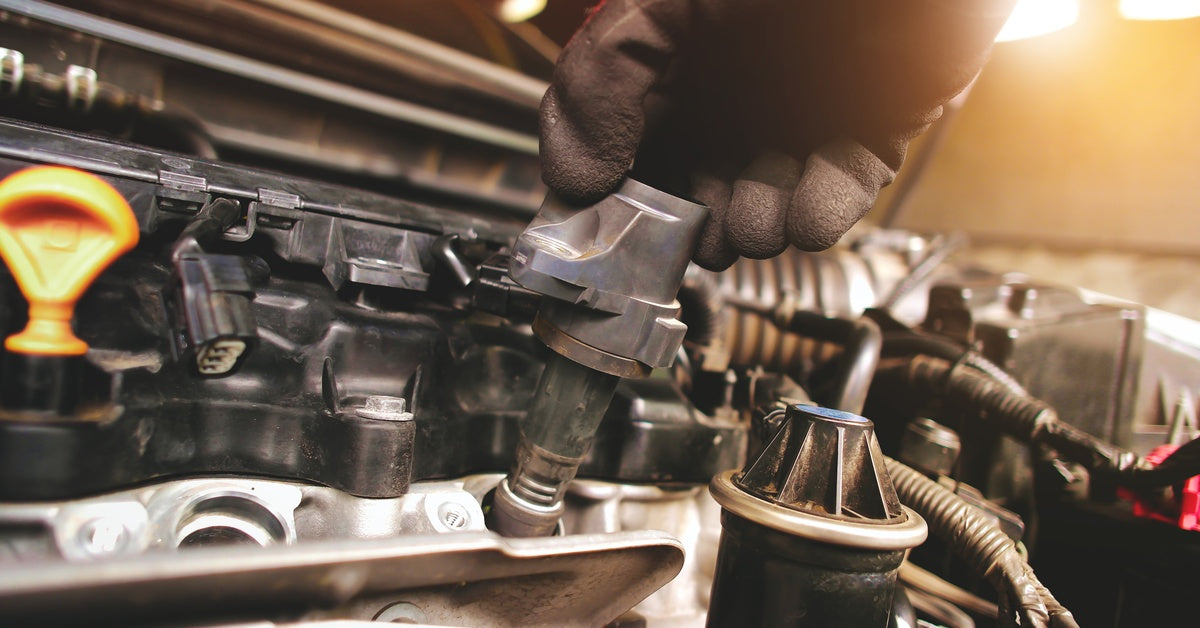 A gloved hand works on a car engine, touching a black ignition coil amid wires, hoses, and an orange cap.