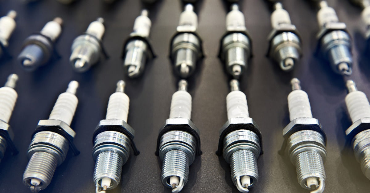 A close-up image of multiple spark plugs arranged in rows of three on a black reflective surface under bright lighting.