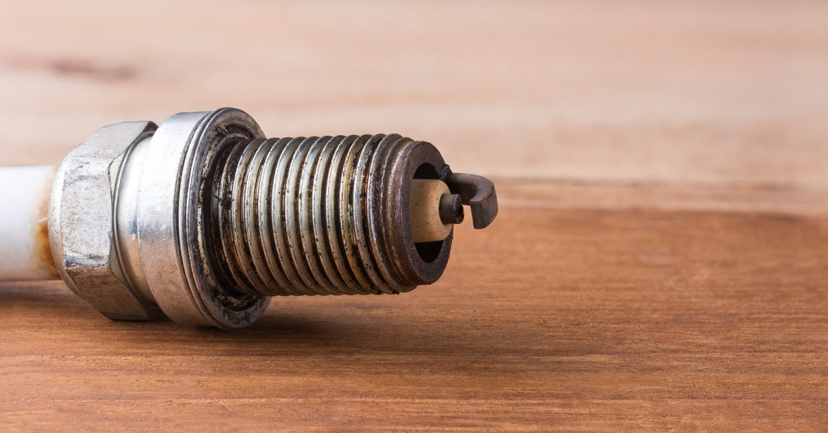 An extreme close-up of a spark plug resting on a wooden surface, showing black residue and a dark electrode tip.