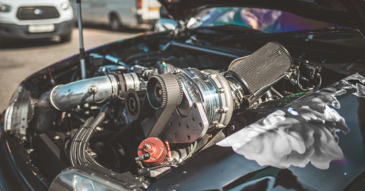 Close-up of a high-performance car engine with exposed supercharger and intake components inside an open hood.