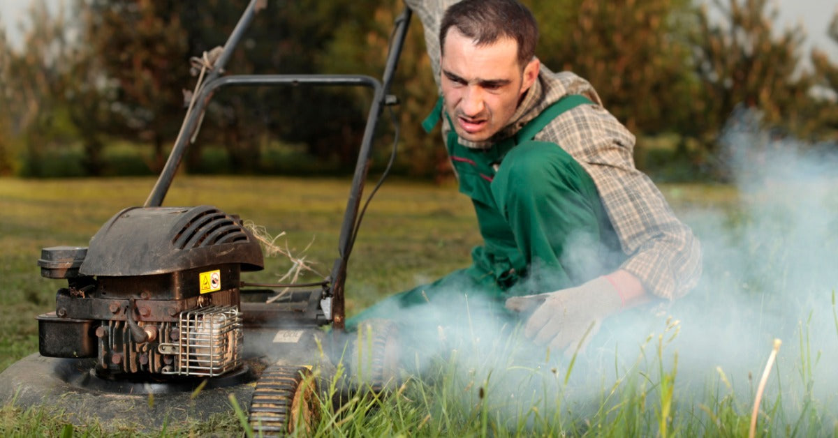 A man in green overalls crouches to inspect a lawn mower engine, which is emitting white smoke outdoors.
