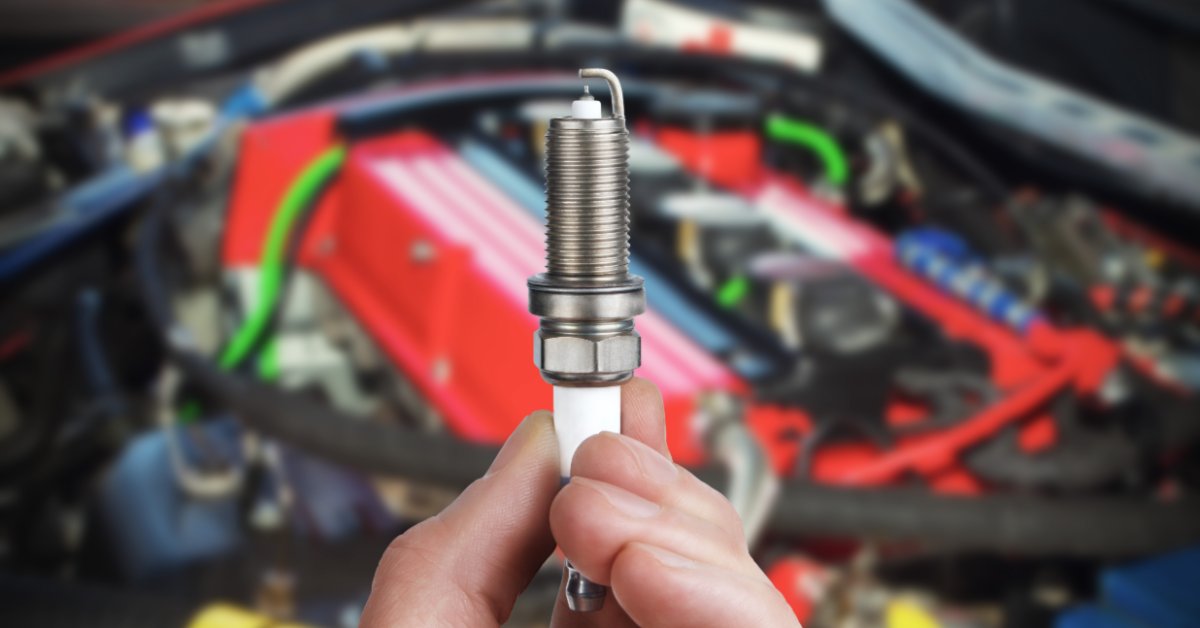 A close-up of a person holding a spark plug, with automotive tools and equipment blurred in the background.