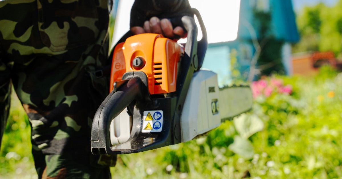 A person wearing camouflage pants is holding an orange chainsaw among green plants in a bright outdoor setting.
