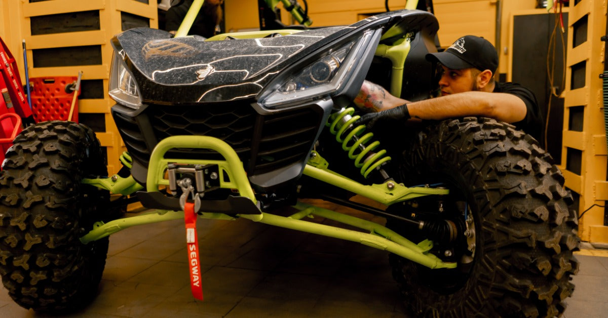 A man inspects an ATV in a workshop with tools hanging on the nearby wall. He reaches behind the vehicle's hood.